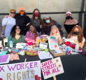 nine swopla members crowd around a table in two rows. the table features condoms, informational pamphlets, and is decorated with protests signs calling for sex worker rights. everyone is smiling at the camera.
