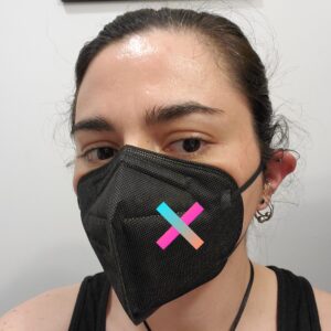 Selfie of me in three-quarter view. I am a white person with dark hair pulled back away from my face. I'm wearing a black tank top and a black KN95 mask with a very obvious cleanly folded line down the middle from the top of my nose to the seam in the front of the mask. A colorful X icon has been pasted in over the left cheek of the mask.